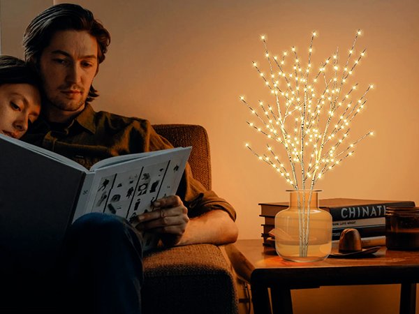 Couple reading a book next to a lighted birch tree in a vase on a table