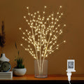 Illuminated birch tree branches with warm white LED lights in a glass vase on a wooden surface, with decorative items on the side.