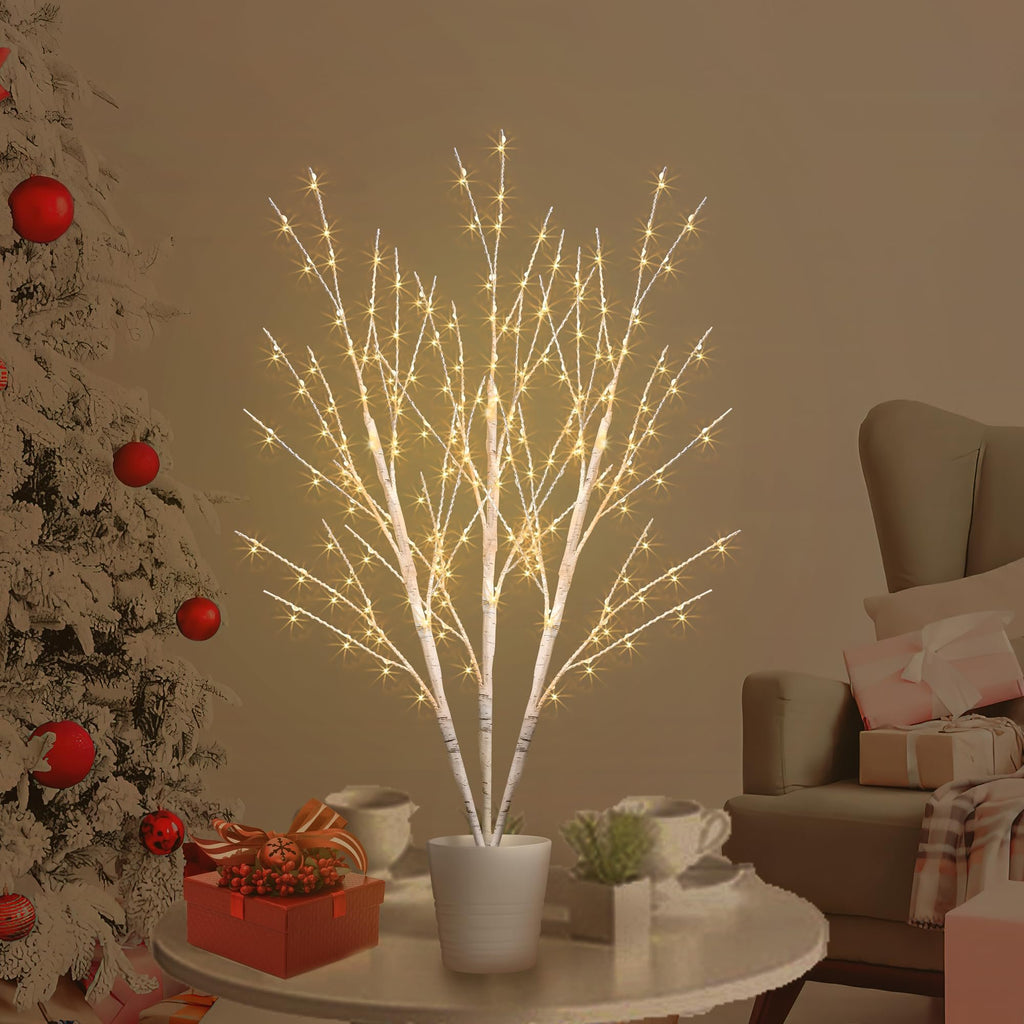 Lighted birch tree with 180 LED lights in a white pot, set on a table with gifts and cups, near a decorated Christmas tree and armchair.