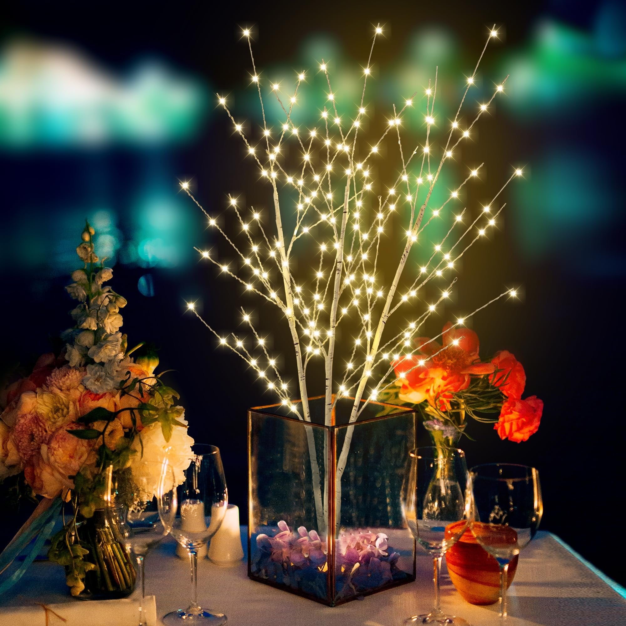 Illuminated birch tree branches with 180 LED lights in a glass vase on a table setting with flowers and wine glasses.