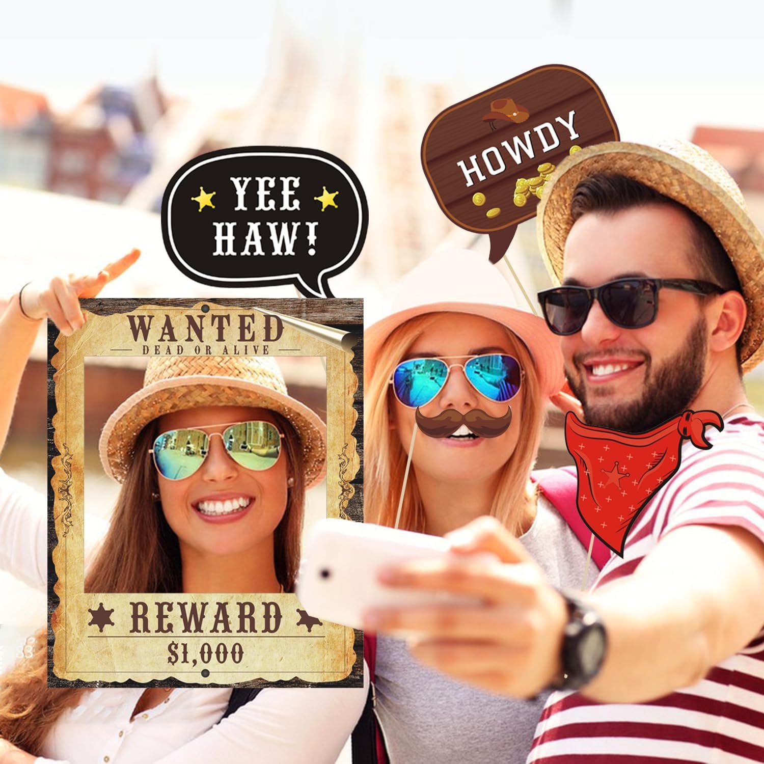 Two smiling people taking a selfie with Western-themed photo booth props including a wanted poster frame, mustache, bandana, and speech bubbles.
