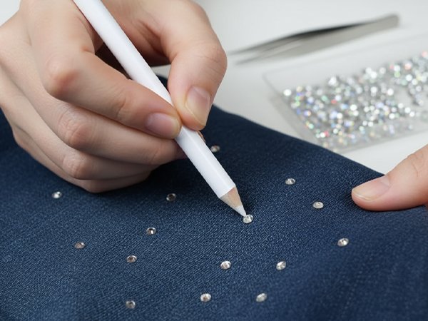 Hands applying clear flatback rhinestones to denim fabric with a white pencil, tweezers and a bag of rhinestones in background.