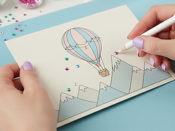 Hands decorating a card with rhinestones and a pencil, featuring a hot air balloon and mountains.