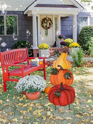 Stacked hand-painted pumpkins garden stake with leaves, placed in front of a red bench and house entrance.