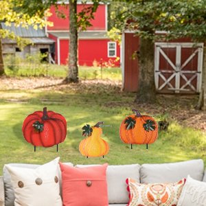 Three stacked metal pumpkins garden stakes with wind-moving leaves on a lawn in front of a red barn.
