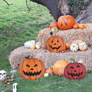 Three carved metal pumpkin garden stakes with Halloween faces, displayed on hay bales with real pumpkins and decorative skulls.