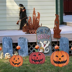 Three jack-o'-lantern garden stakes with pumpkins and skulls in front of a witch decoration and tombstone yard signs.