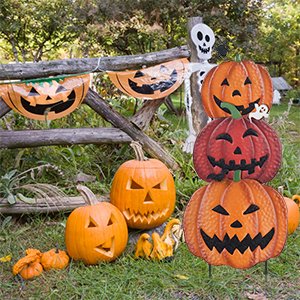 Stackable metal pumpkin garden stakes with carved jack-o'-lantern faces, displayed outdoors near a wooden fence with Halloween decorations.