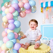 Smiling baby in a white shirt and grey shorts sits next to a pastel rainbow balloon arch with gold confetti balloons.
