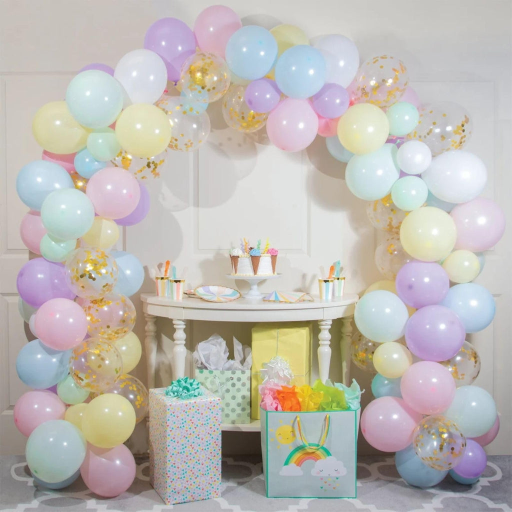 Pastel rainbow balloon arch with gold confetti balloons over a table with a cake and gifts