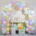 Pastel rainbow balloon arch with gold confetti balloons over a table with a cake and gifts