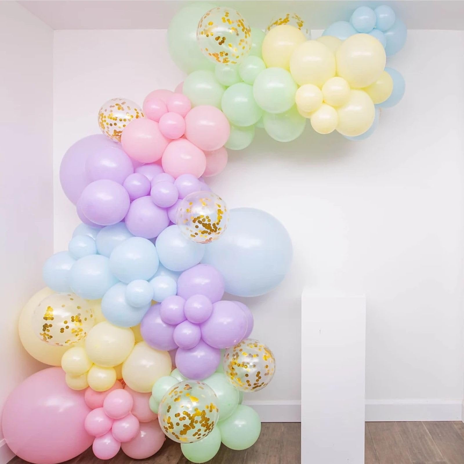Pastel rainbow balloon garland with gold confetti balloons against a white wall and wooden floor