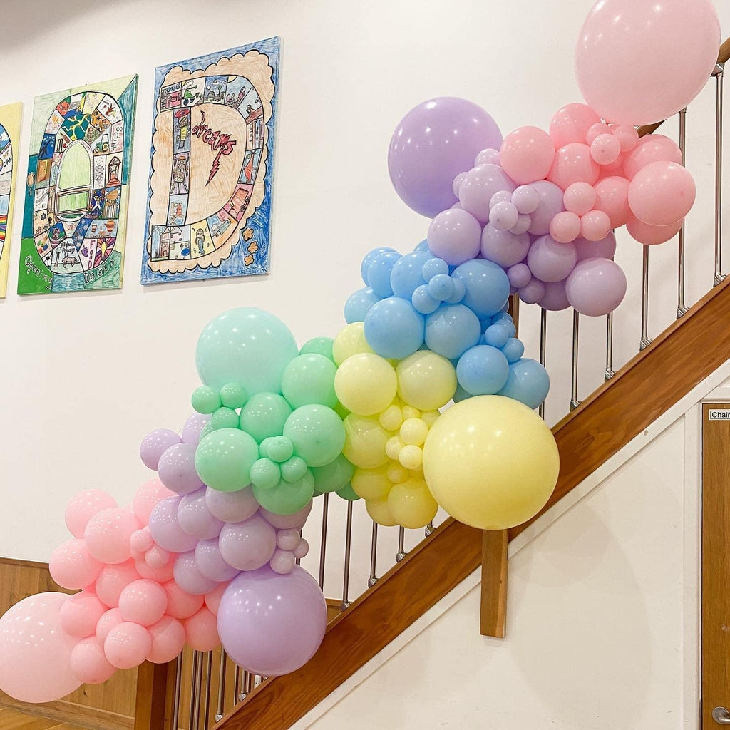Pastel rainbow balloon garland draped over a wooden staircase railing, featuring pink, purple, blue, green, and yellow balloons.