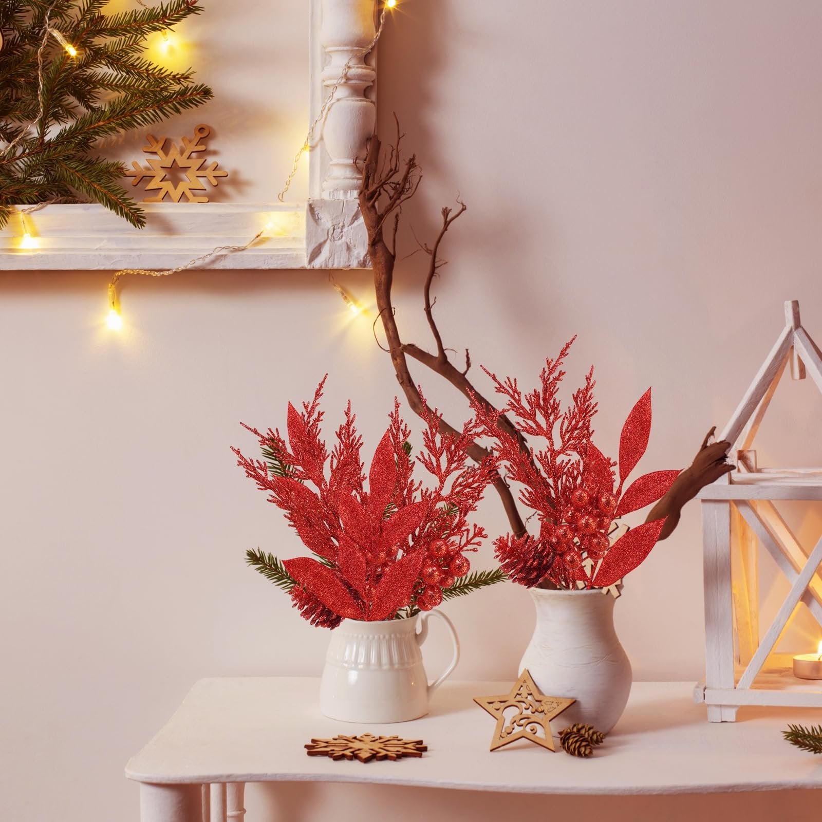 Two white vases holding red glitter poinsettia branches and pine sprigs on a white table with Christmas decor.