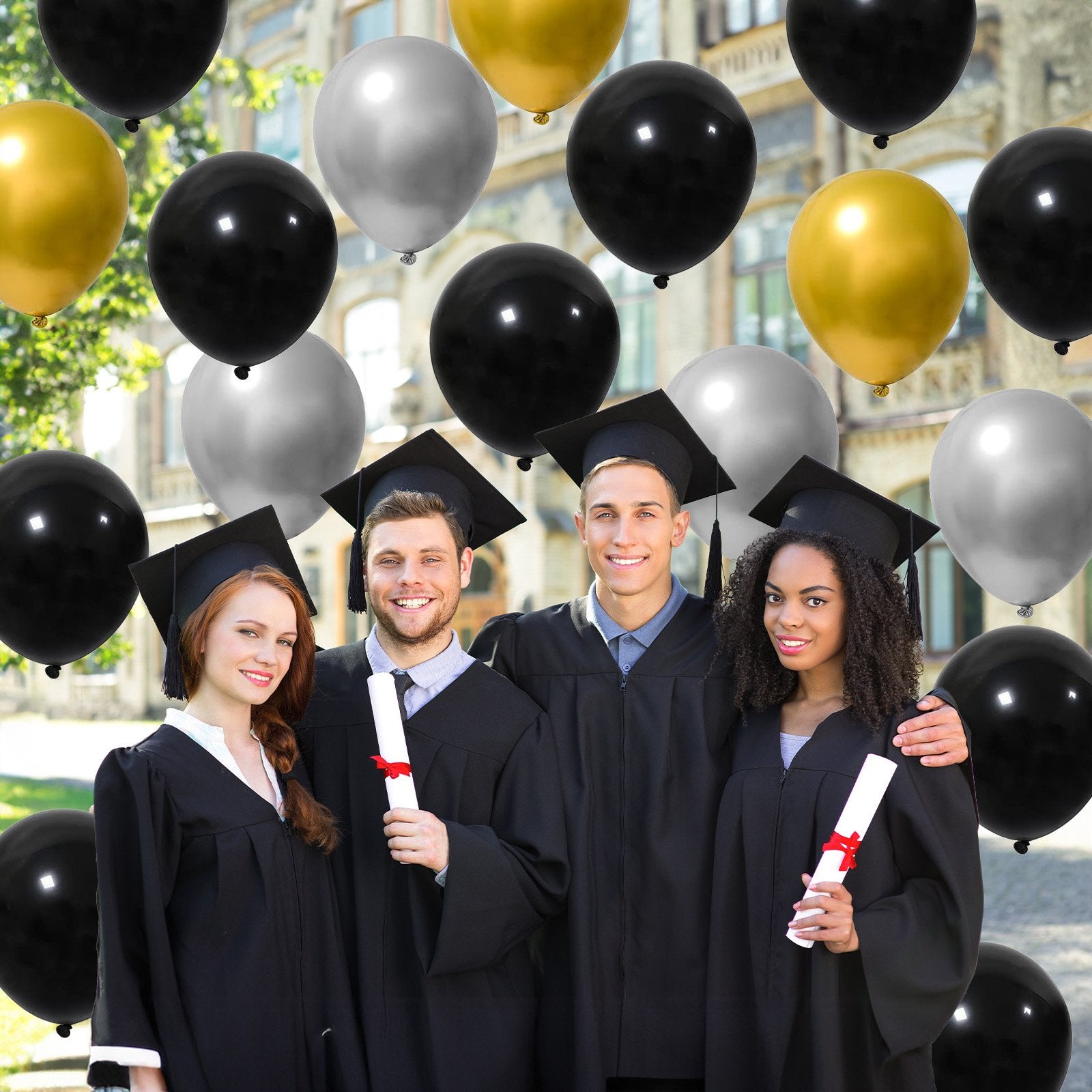 Four smiling graduates in caps and gowns holding diplomas, surrounded by black, gold, and silver balloons.