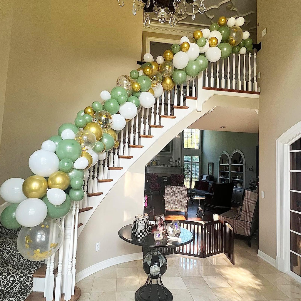 Balloon garland in sage green, gold, and white draped along a staircase railing and wall.
