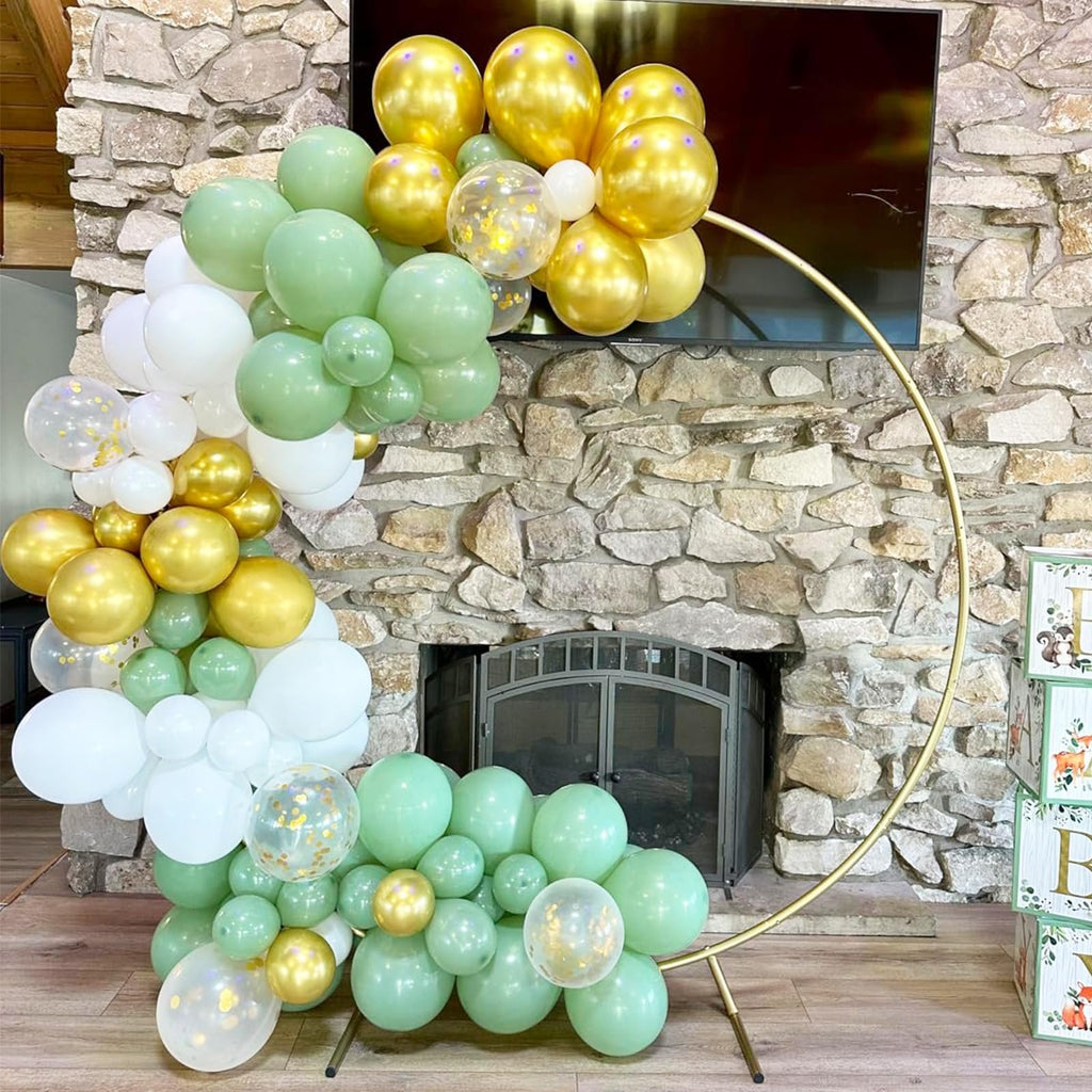 Balloon arch in sage green, gold, white, and confetti balloons arranged around a gold hoop in front of a stone fireplace.