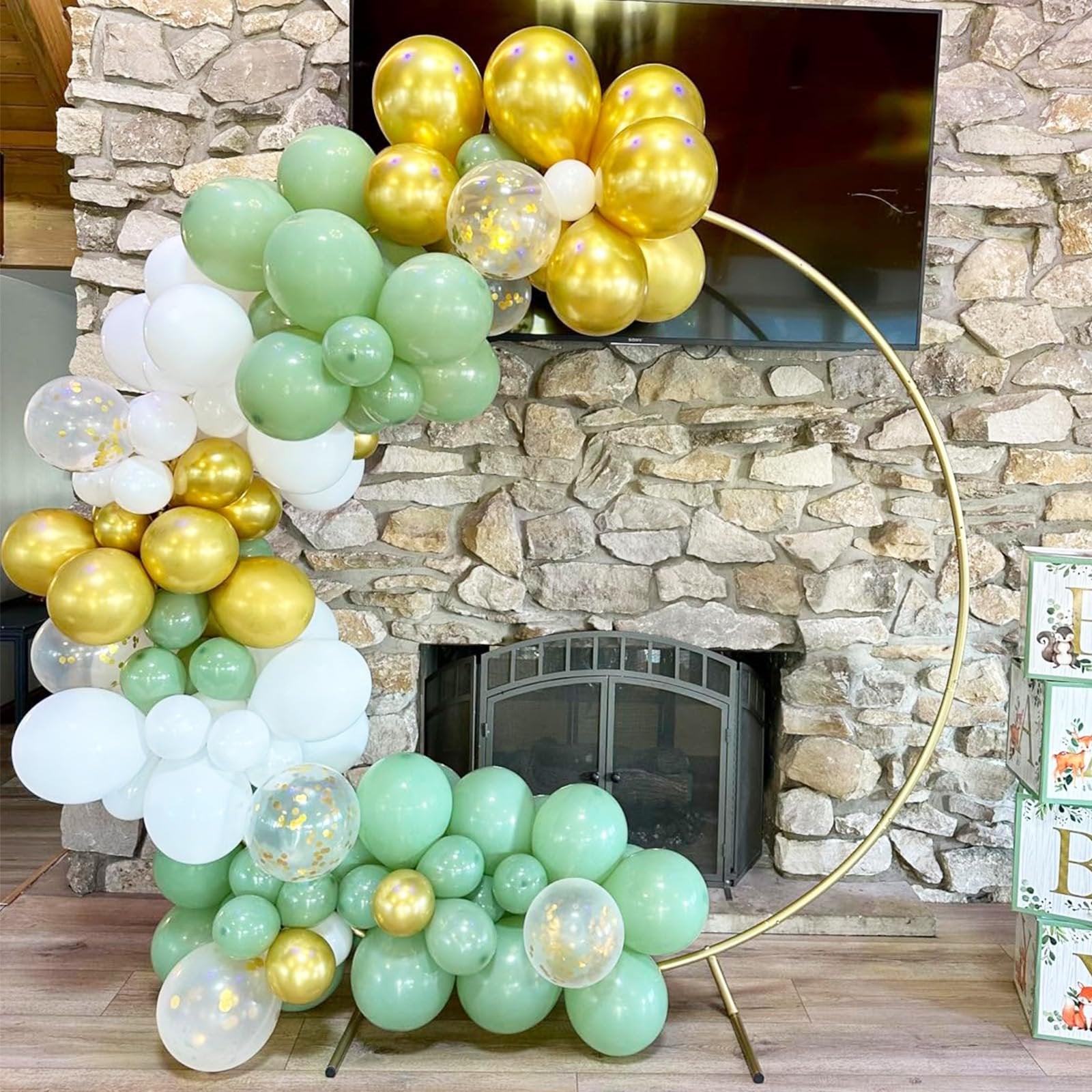 Balloon arch in sage green, gold, white, and confetti balloons arranged around a gold hoop in front of a stone fireplace.