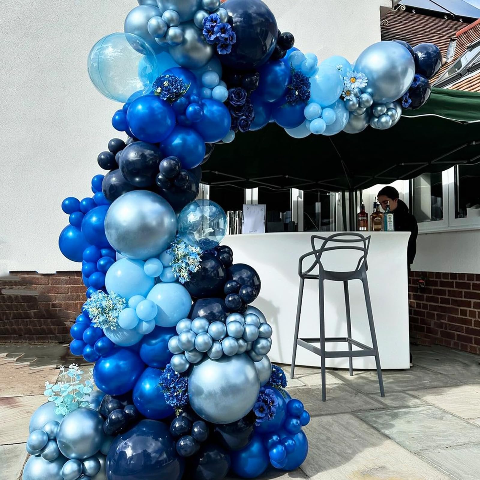 Blue and silver balloon arch with floral accents, set up outdoors near a white bar with a stool.
