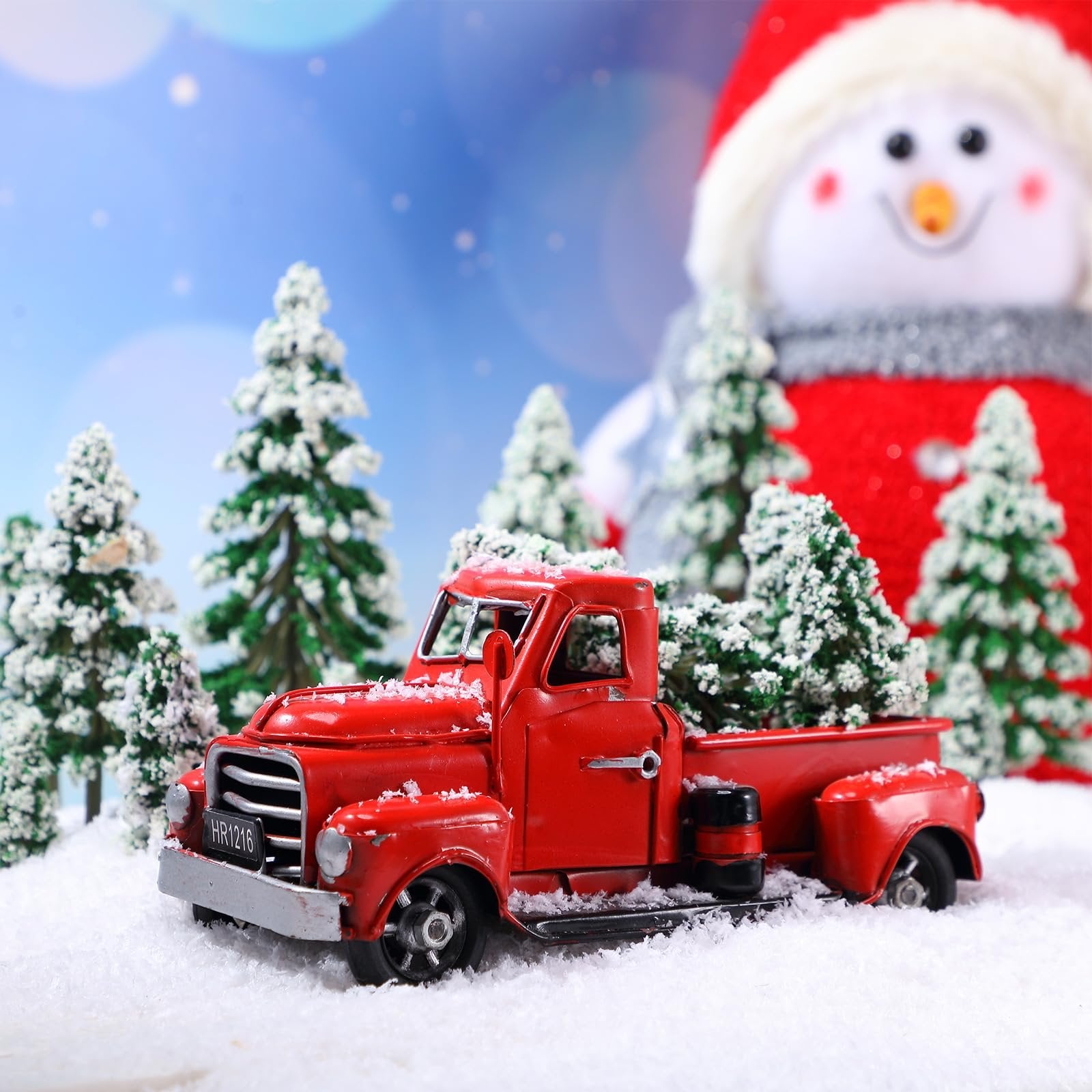 Red vintage truck carrying miniature snow-covered pine trees in a winter scene with a snowman in the background.