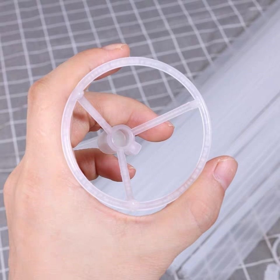 Hand holding a clear plastic balloon holder with three spokes and a central hub, against a grid background.