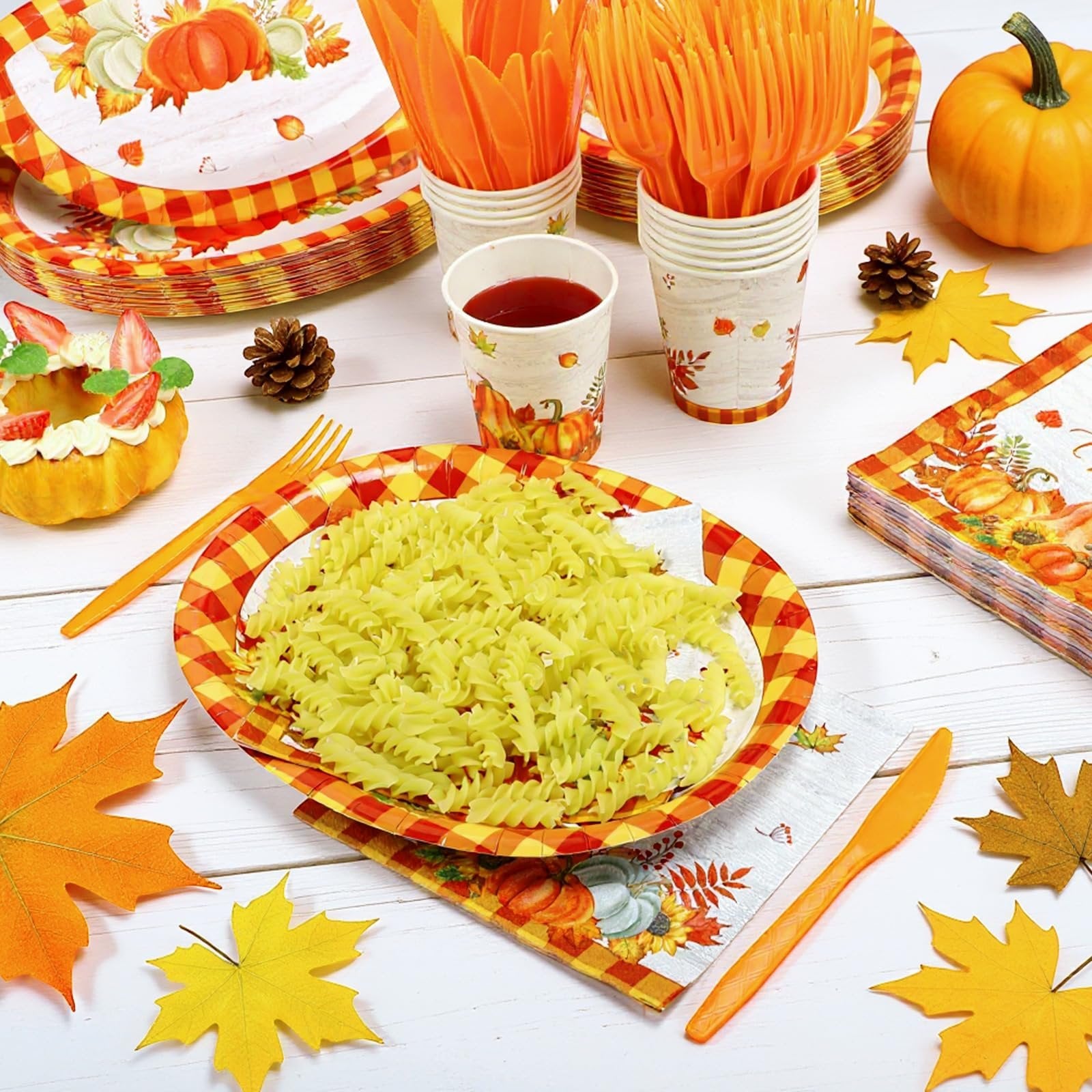 Thanksgiving tableware set with pumpkin and plaid design, featuring plates, cups, napkins, and cutlery, arranged on a white wooden table with autumn leaves and a small pumpkin.