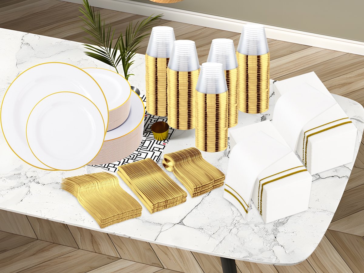 White and gold disposable dinnerware set including plates, cups, napkins, and cutlery arranged on a marble table.