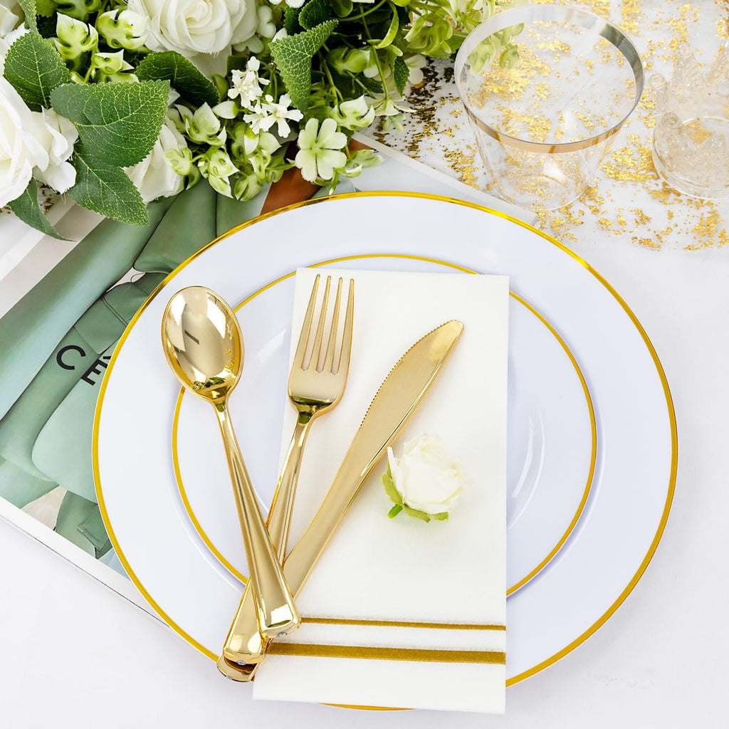 White and gold disposable dinnerware set with cutlery and napkin on a plate, with floral arrangement and cups in background.
