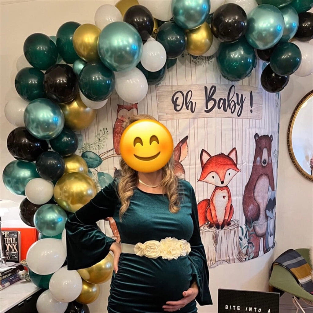 Pregnant person in green dress with balloon arch and woodland creature backdrop