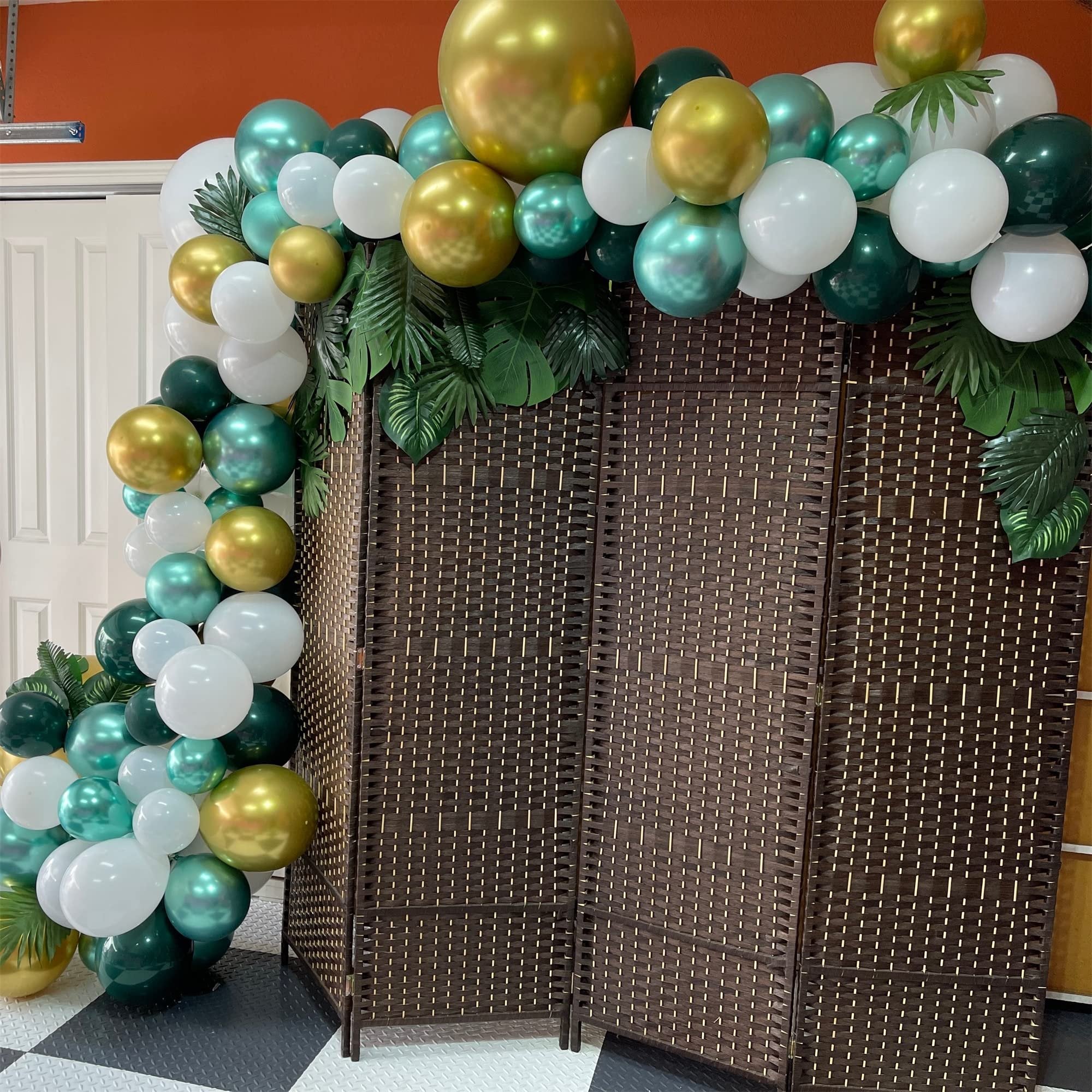 Balloon garland in dark green, gold, and white with tropical leaves draped over a woven screen.