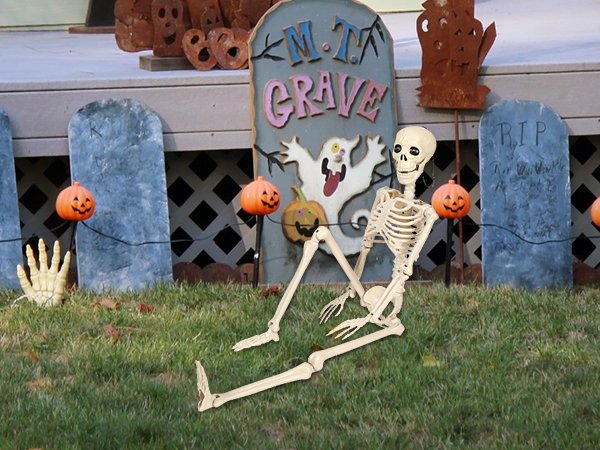 Poseable skeleton prop sitting on grass in front of Halloween tombstone decorations and pumpkins