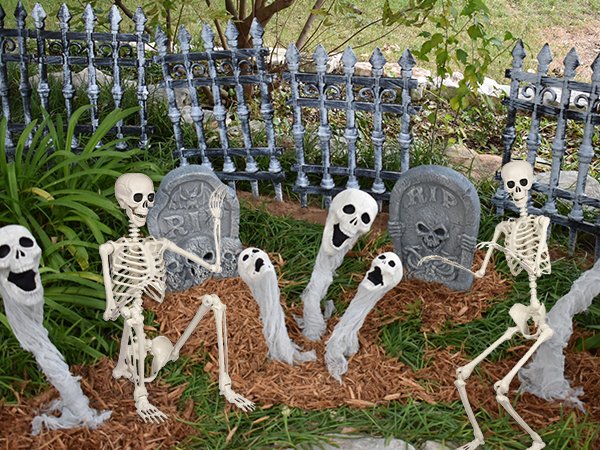 Group of Halloween skeletons and ghosts in a graveyard setting with tombstones and a fence.