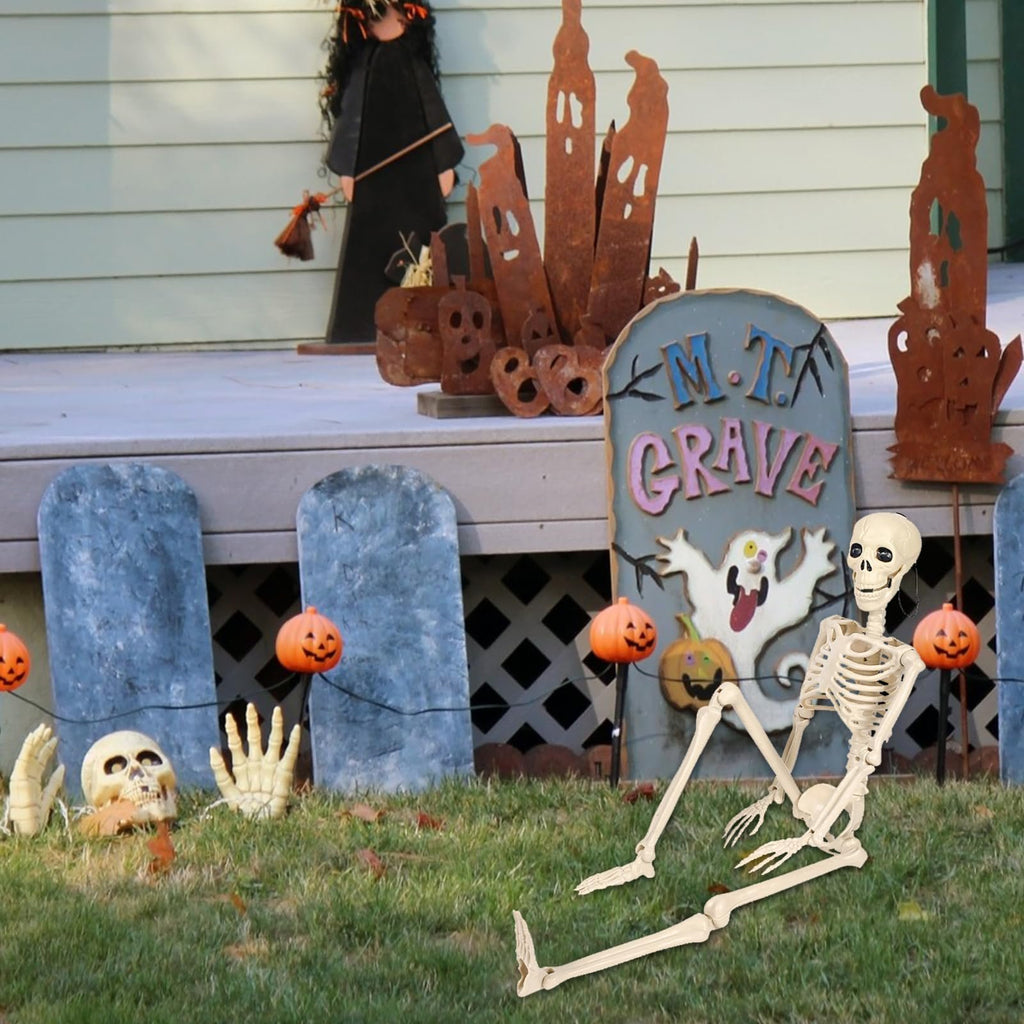 Poseable skeleton prop sitting on grass in front of Halloween decorations including tombstones and pumpkins.