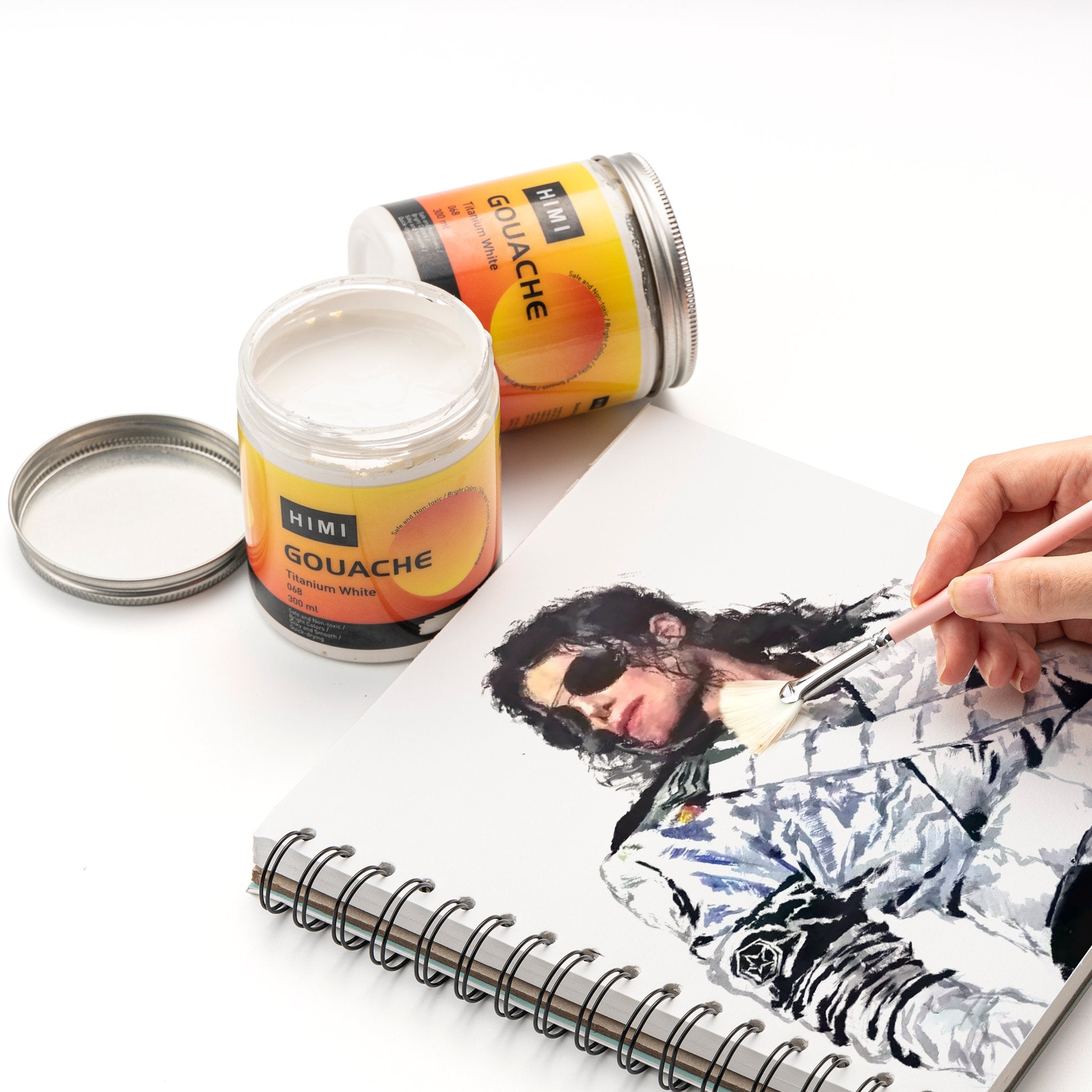 Two jars of white gouache paint and a sketchbook with a portrait being painted by a hand holding a brush.