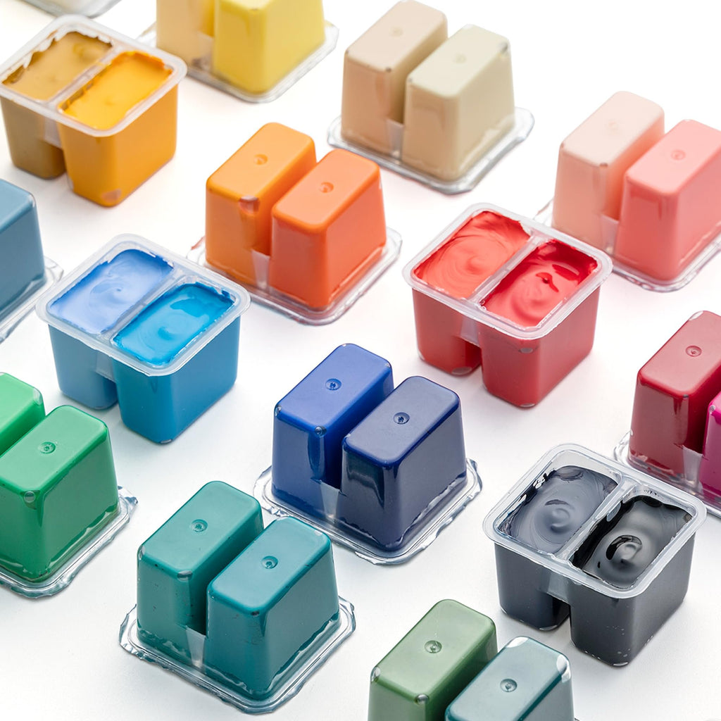 Assortment of twin-cup gouache paint pots in various colors, arranged on a white surface.