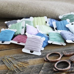 Embroidery floss bobbins with thread, needles, and scissors on a wooden surface with burlap background.