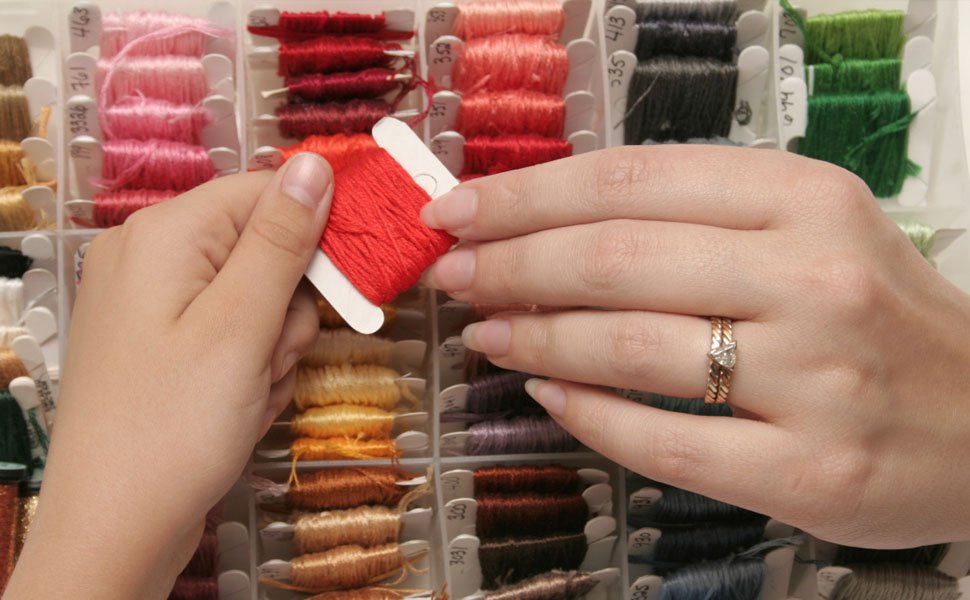 Hands organizing red embroidery floss onto a plastic bobbin from a compartment organizer box filled with colorful threads.