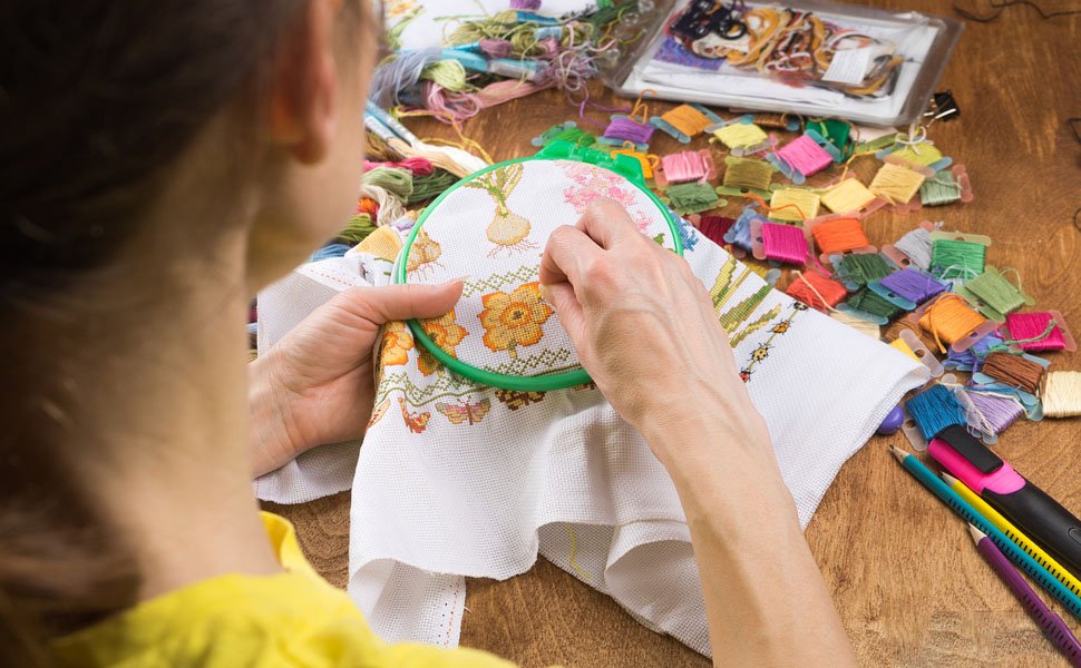 Person's hands embroidering a floral pattern on white fabric held in a green hoop, surrounded by colorful thread spools.