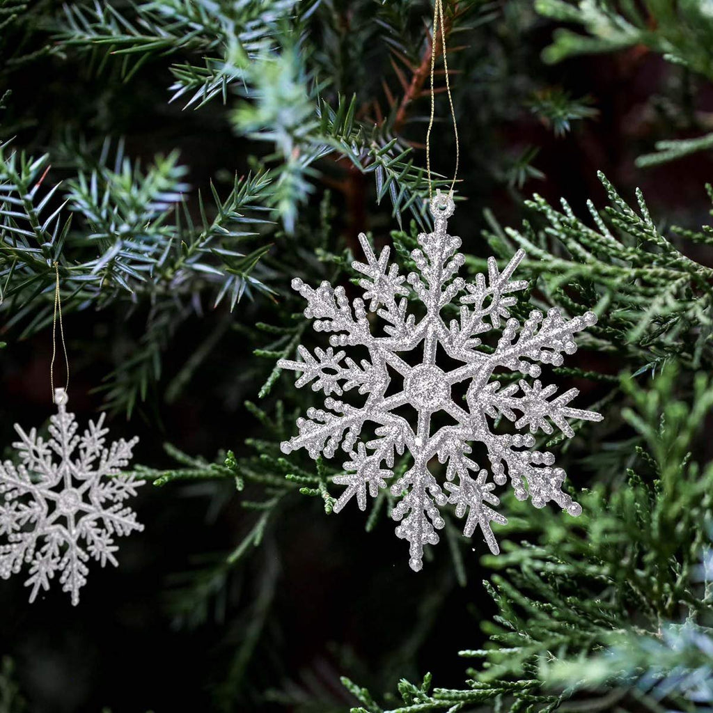 Two silver glitter snowflake ornaments hanging on a Christmas tree branch, one in focus and larger, the other smaller and slightly out of focus.