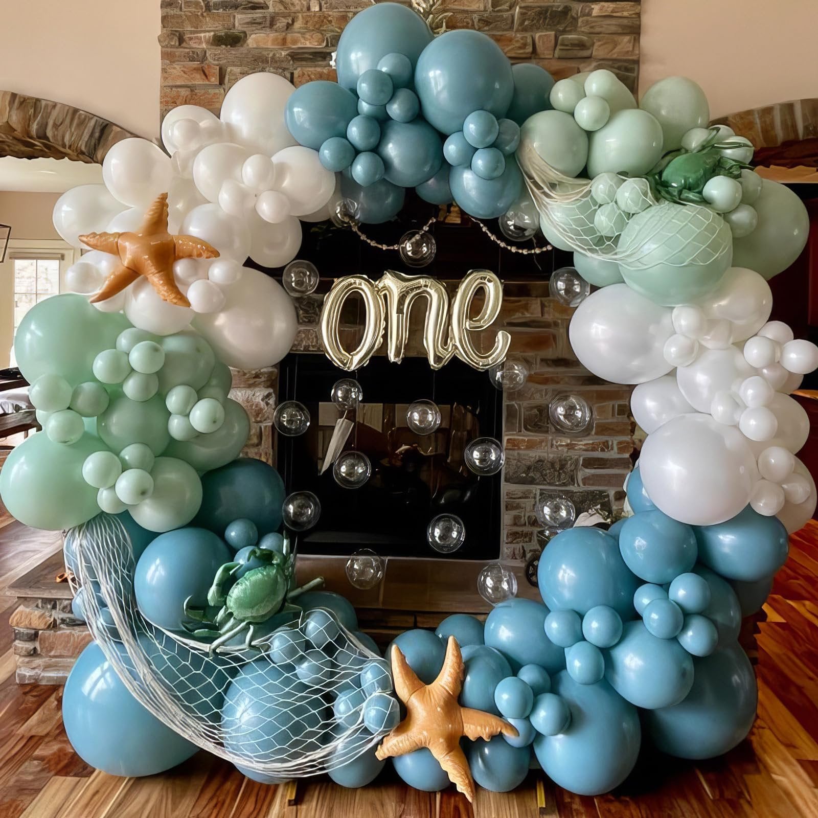 Balloon garland with starfish and crab decorations in blue, green, white, and gold, spelling out 'one' in front of a fireplace.