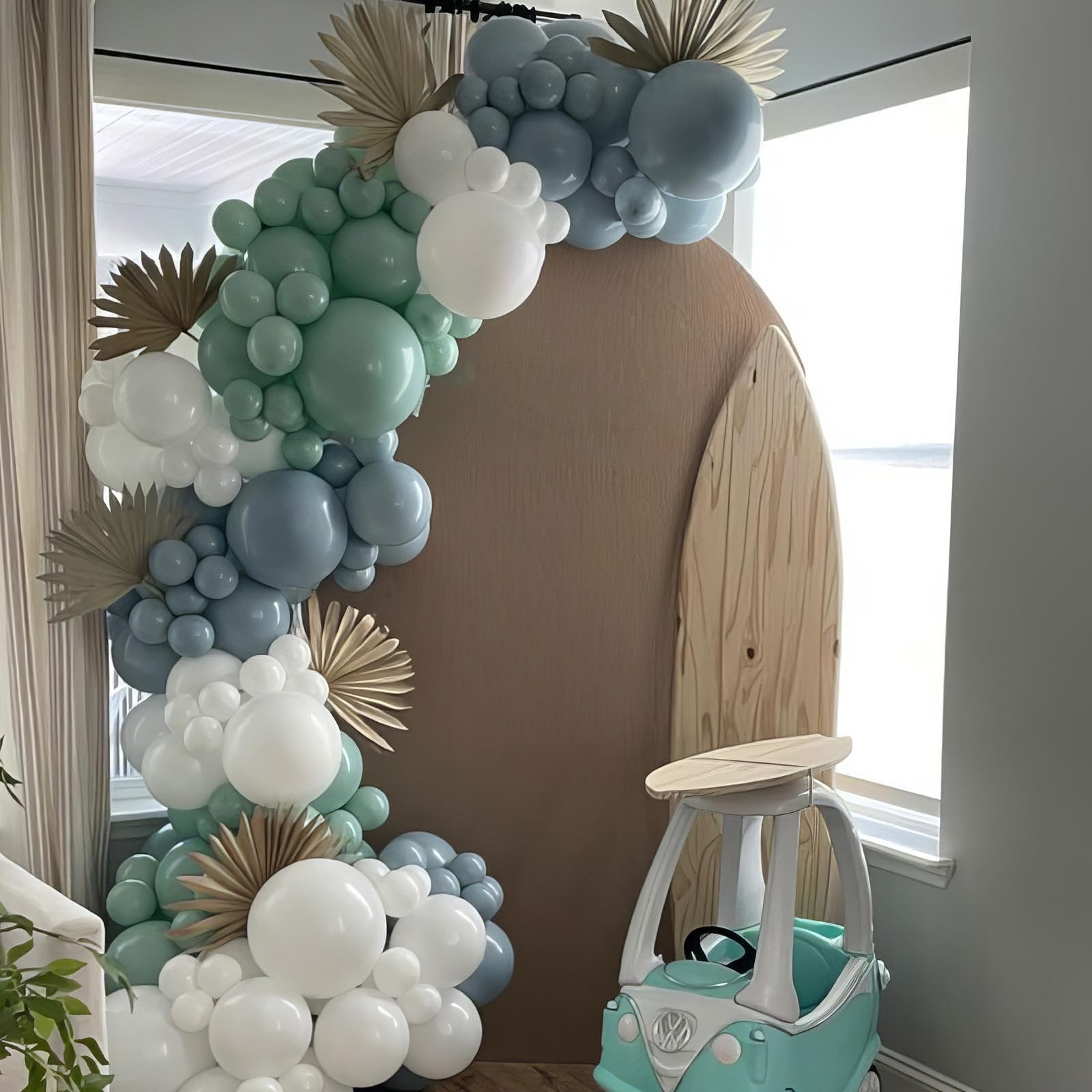 Balloon arch in dusty blue, sage green, white, and gold with dried palm leaves, next to a wooden surfboard and toy car.