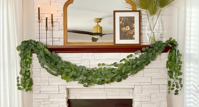 Artificial eucalyptus garland draped over a white stone fireplace mantel with candles and artwork.