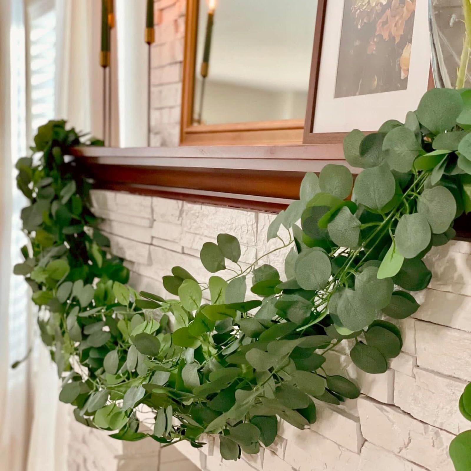 Artificial eucalyptus garland draped over a white brick fireplace mantel with candles and a framed picture in the background.