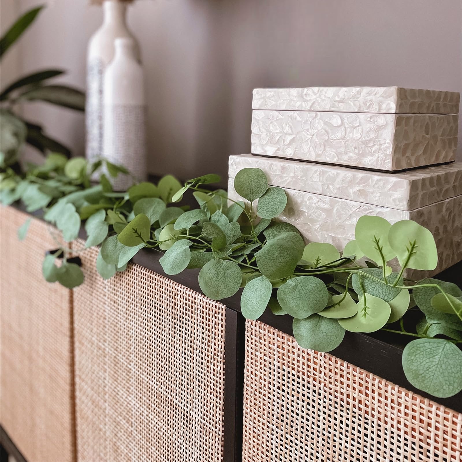 Artificial eucalyptus garland draped over a cane-front cabinet with decorative boxes and vases.