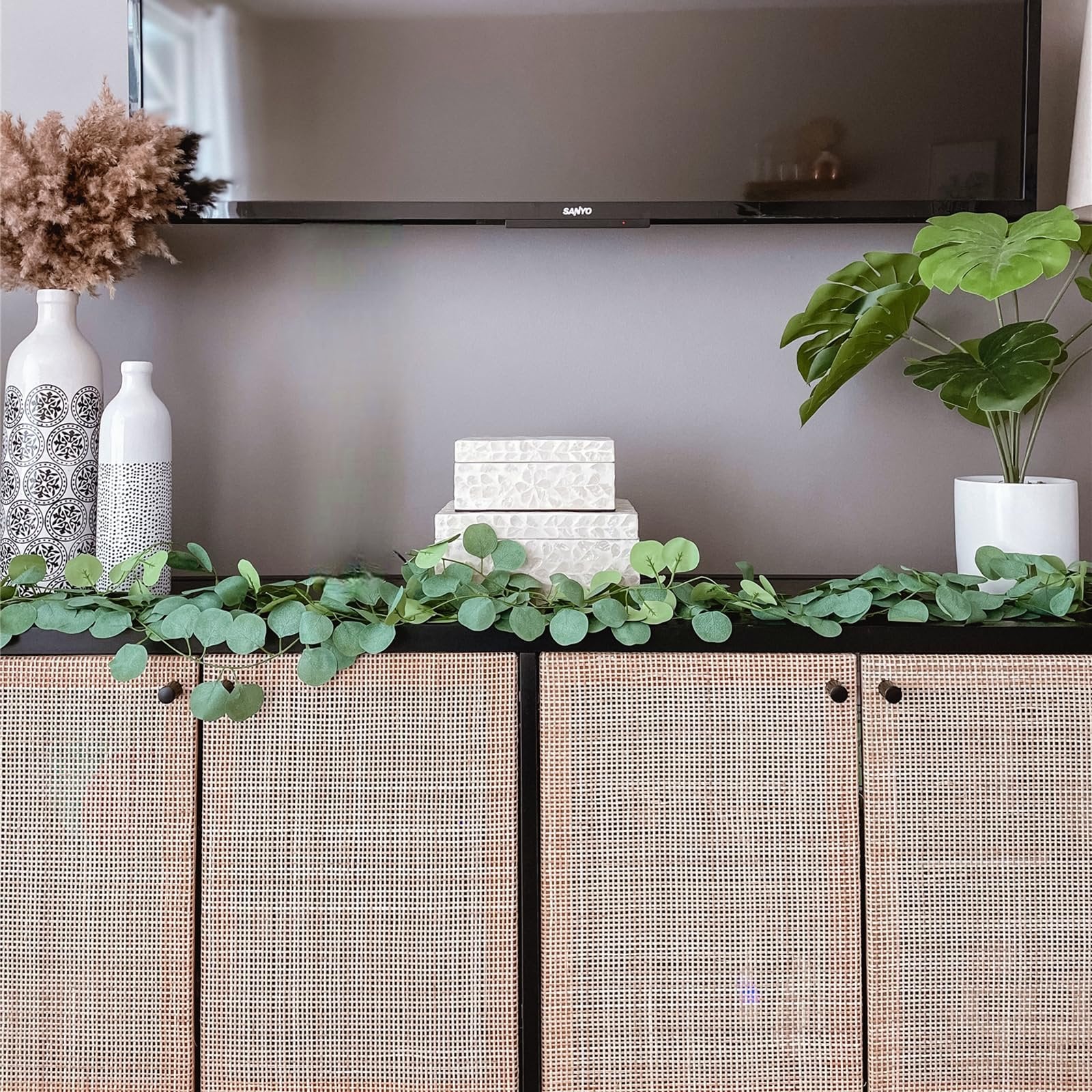 Artificial eucalyptus garland draped across a black cabinet with woven doors, flanked by decorative vases and boxes.