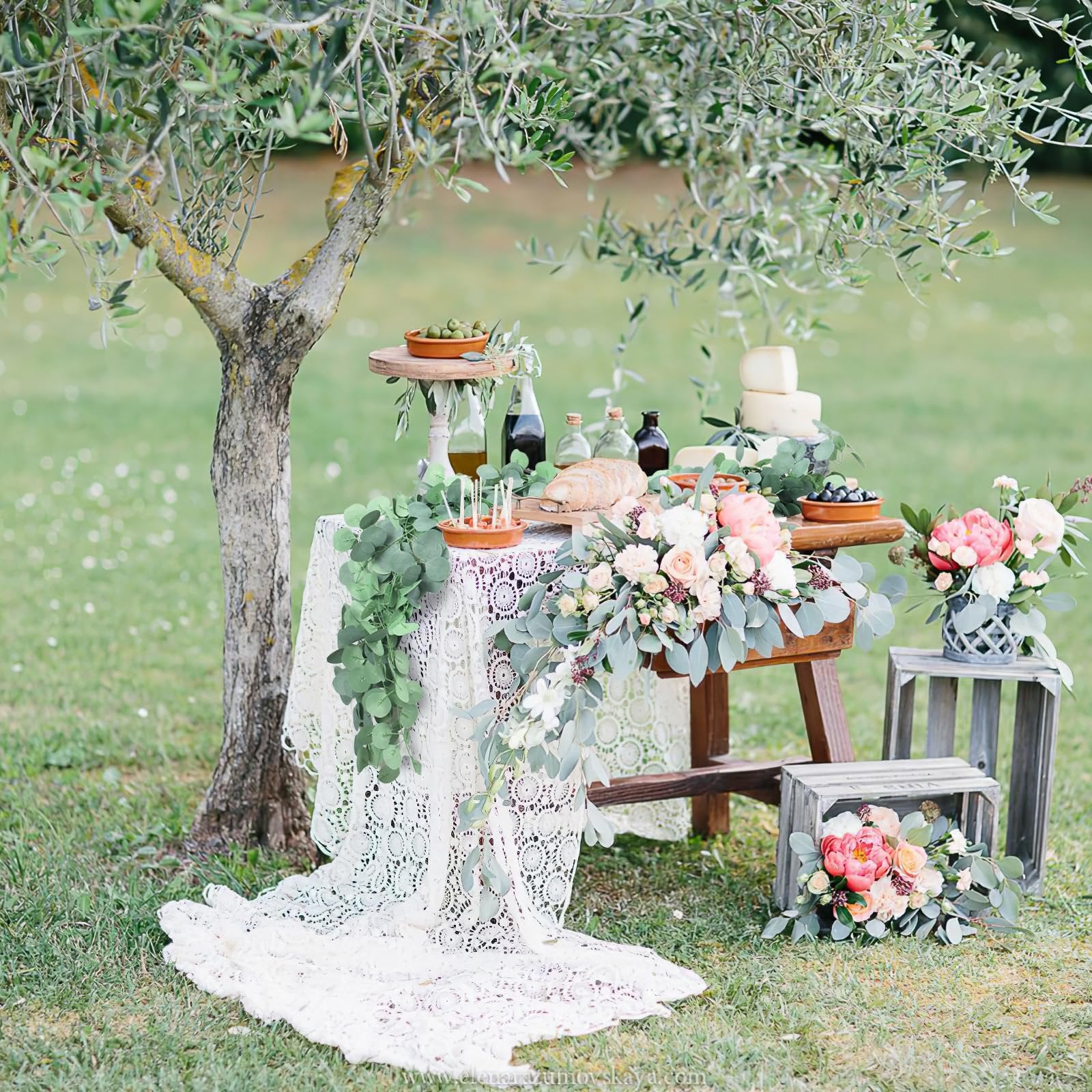 Outdoor table setting with artificial eucalyptus garland, flowers, bread, cheese, and olives under an olive tree.