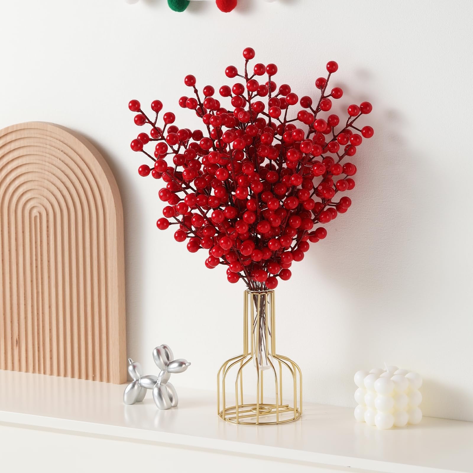 Bunch of artificial red berry stems in a gold wire vase on a white shelf, with decorative items.