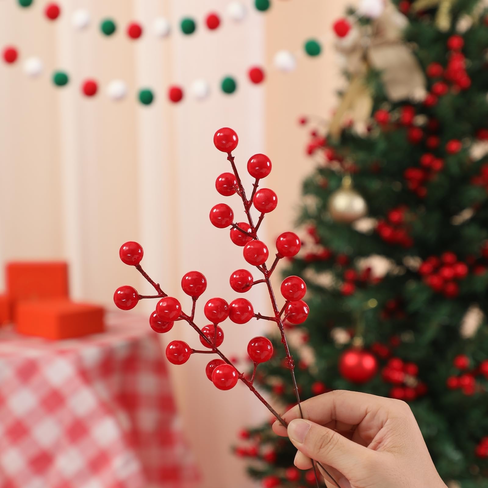 Hand holding artificial red berry stems with a blurred Christmas tree and red gift boxes in the background