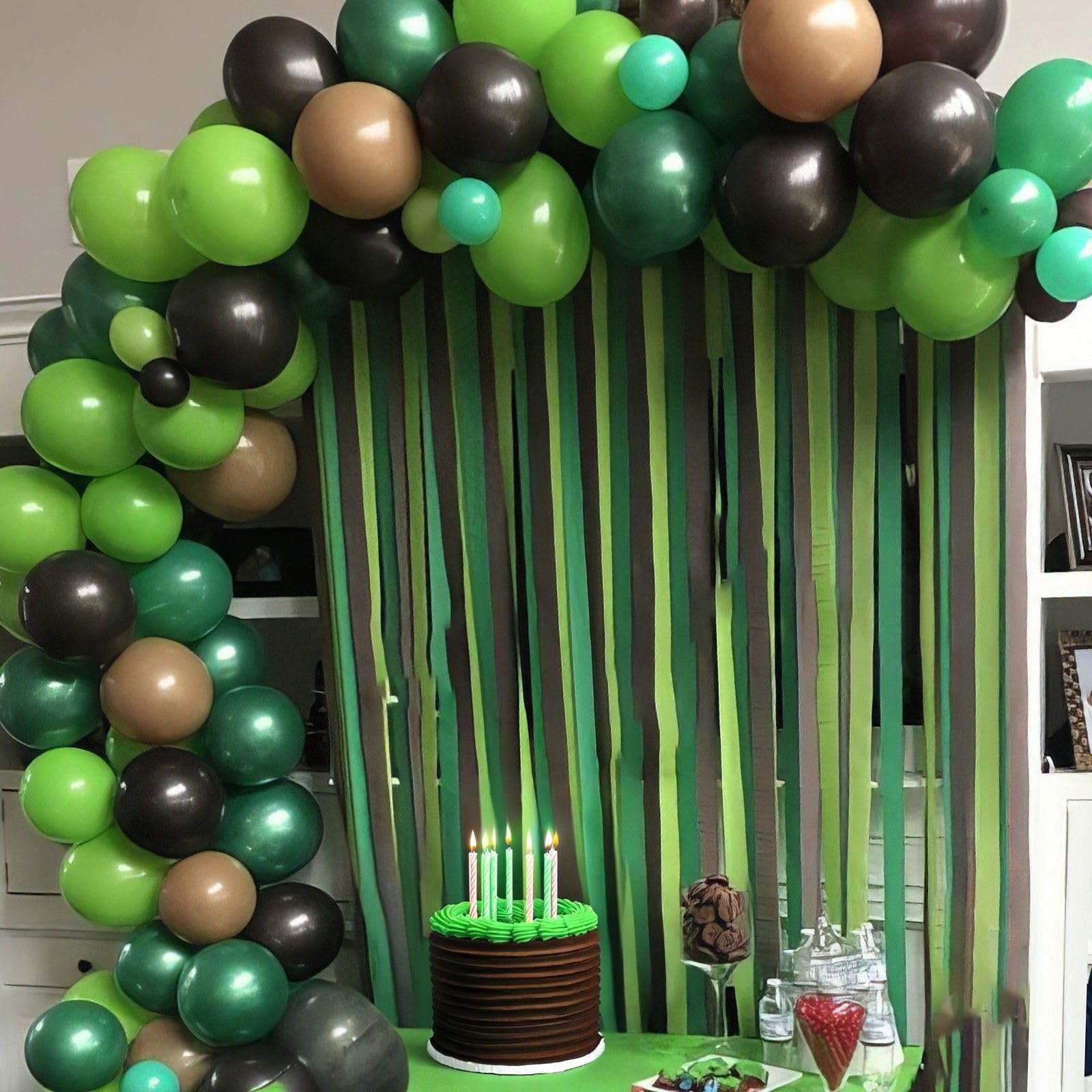 Camouflage-themed birthday party setup with a balloon arch, striped backdrop, and a chocolate cake with green frosting and candles.