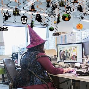 Woman in witch hat and costume at office desk with Halloween swirl decorations hanging from ceiling